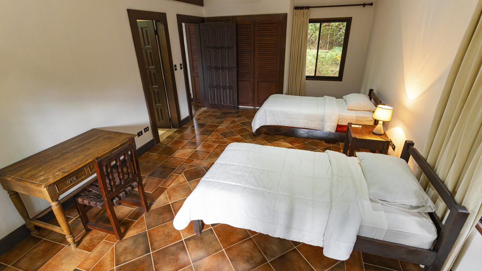 A plain twin room with white bedding, a wooden desk and a terracotta-tiled floor