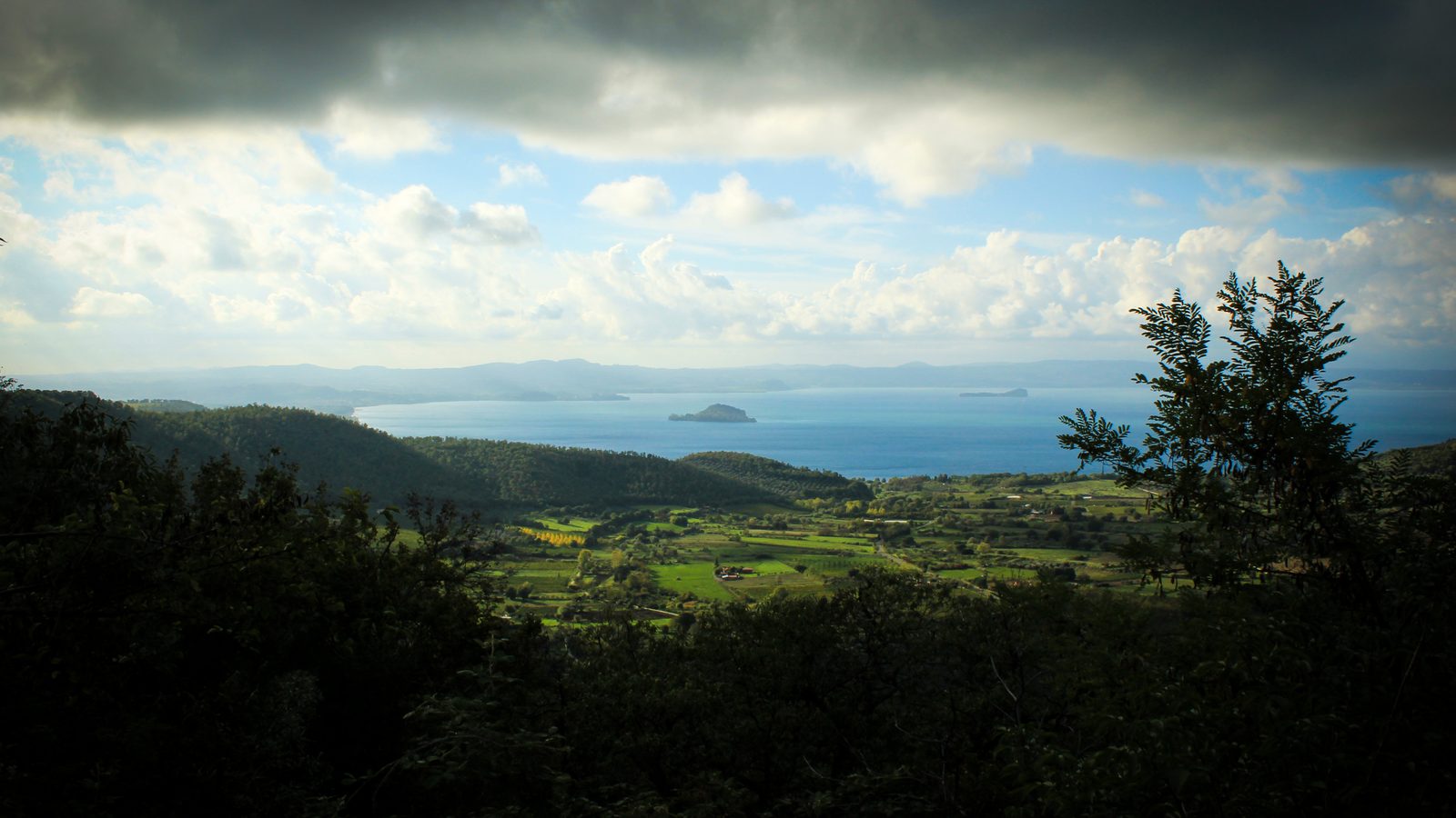 Lake Bolsena seen from wooded hills above its shore, with clouds over the water